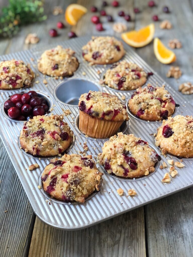 Ten baked muffins in muffin tin, one popping up, cranberries in one muffin cup, oranges, walnuts surrounding.