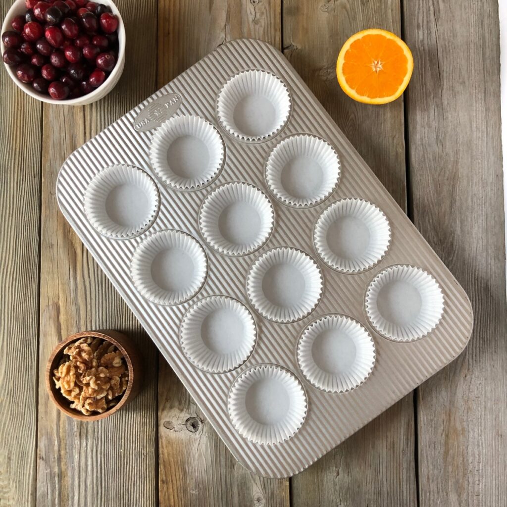 Muffin tin prepared with paper muffin liners on a wood board with orange, cranberry walnuts near it.