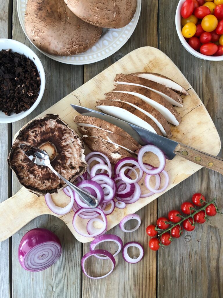 Mushrooms and onions being sliced on a wood board.