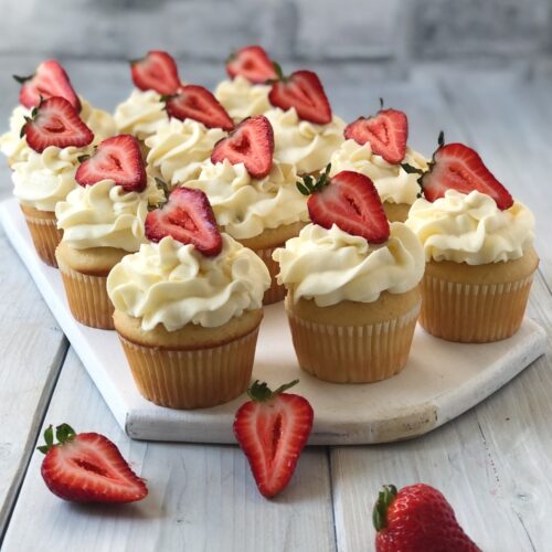 Twelve frosted strawberry cupcakes on a white board garnished with sliced strawberries.